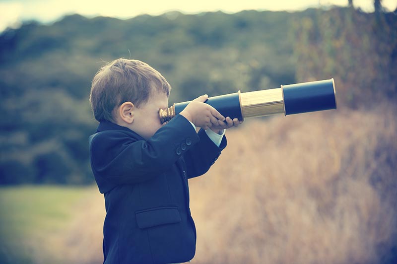 Young boy in a business suit with telescope. Small child wearing a full suit and holding a telescope. He is holding the telescope up to his eye. Business forecasting, innovation, leadership and planning concept. Shot outdoors with trees and grass in the background