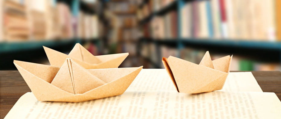 Photo of two origami boats sitting on books in the foreground of a library