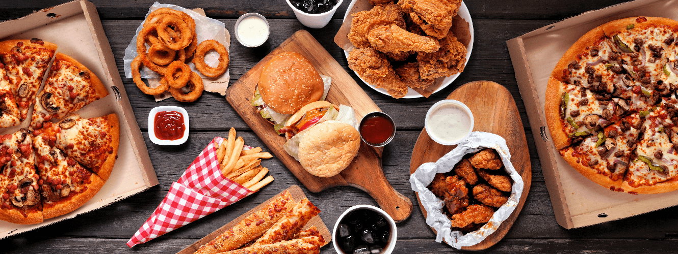 Table of assorted take out or delivery foods. Hamburgers, pizza, fried chicken and sides. Top down view on a dark wood banner background.